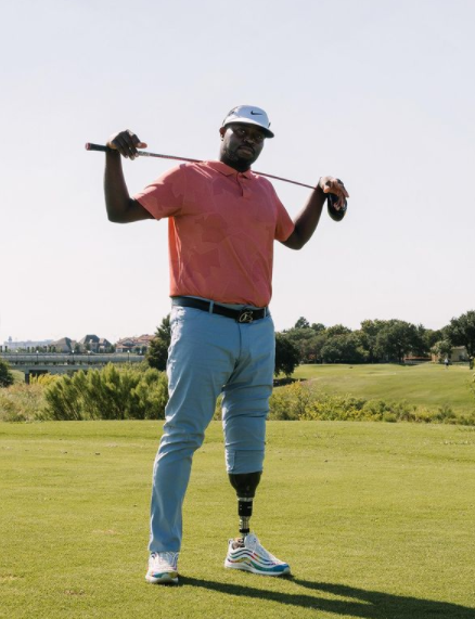 This image shows golfer Carlos Brown stood with a golf club over his shoulders and both hands resting on it. He is wearing a white cap white a black Nike tick, a coral polo shirt, light blue trousers with a black belt, and white and blue striped AirMax 97s. On his left leg the trousers are rolled up to the knee, revealing his black prosthetic leg. He is stood on a golf course, in the background of the image there is green grass and a clear sky.