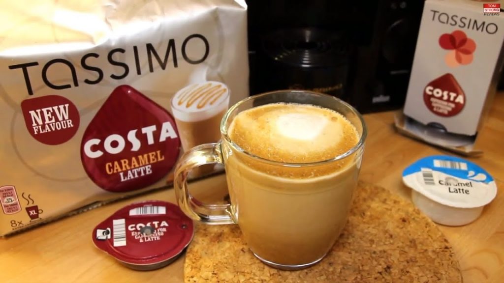 Image shows a light wooden kitchen surface. On it there is a packet of Costa caramel latte Tassimo podss. The coffee and milk pods used are displayed next to a glass mug filled with coffee.