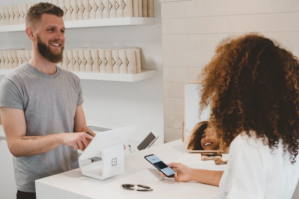 This image shows a bearded man wearing a grey shirt working as a cashier. He is stood behind a white desk  using a small white tablet. He is serving a woman with dark curly hair in a white shirt who is using her phone to pay. In the background of the image there is white shelving lined with products.